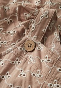 Dulkių rožinė medžiaga su išsamia balta gėlių išsiuvinėta raštu. Medinis mygtukas su gėlių raštu užsegamas drabužį. Lygus tekstūras.