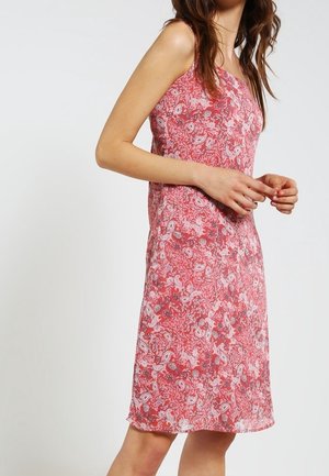 Femme portant une robe rose sans manches, longueur genou, avec un motif floral blanc et rouge, debout devant un fond clair uni.