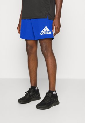 Niebieskie spodenki sportowe z białym logo Adidas obok czarnych butów sportowych i skarpet, noszone na nogach osoby na jasnym tle.