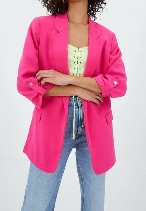 Vrouw met gekruiste armen in een helderroze blazer over een lichtgroene vetershirt en high-waisted blauwe jeans voor een witte achtergrond.