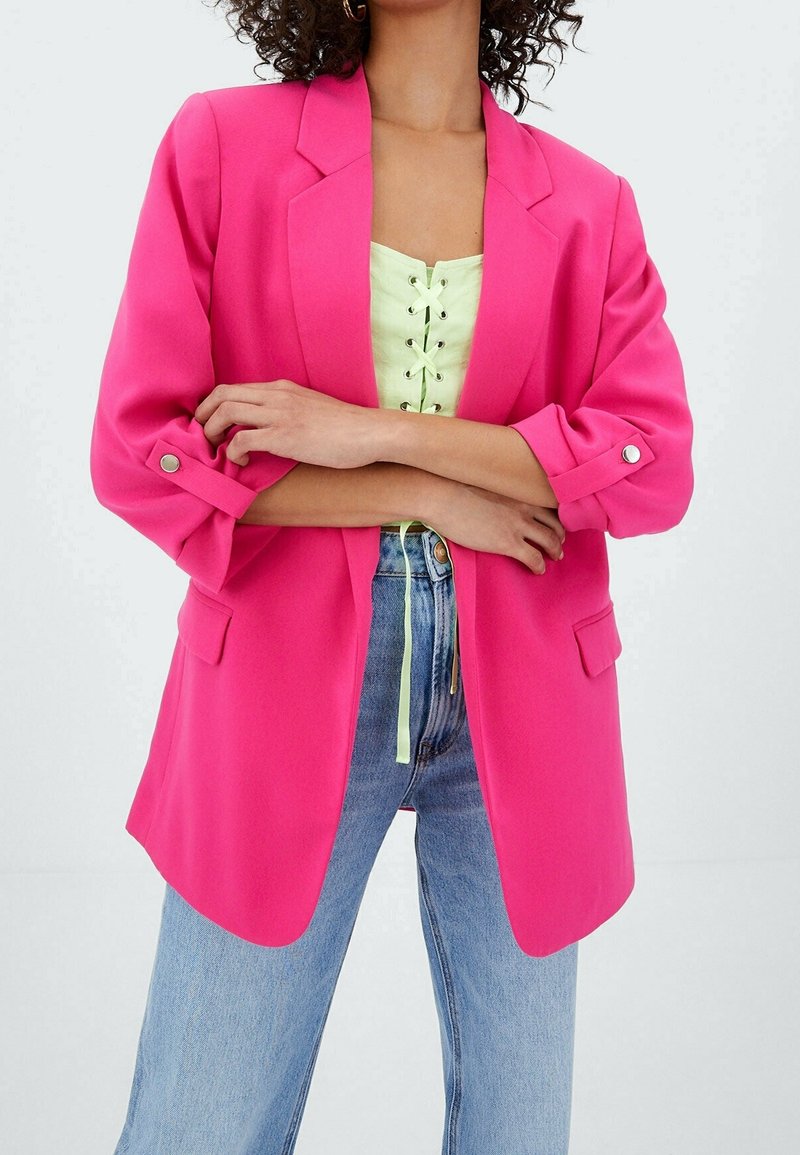 Mujer con los brazos cruzados que lleva puesta una blazer rosa brillante sobre una blusa verde clara con cordones y jeans de tiro alto azul, frente a un fondo blanco.