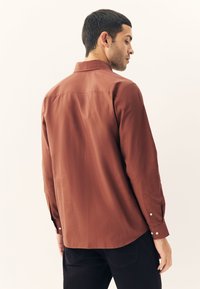 Chemise à manches longues de couleur rouille, fabriquée en tissu lisse. Elle présente un col classique, des poignets à boutons et un ourlet légèrement incurvé à l'arrière.