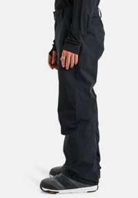 Schwarze wasserdichte Hosen mit geradem Schnitt, zwei seitlichen Taschen und verstellbaren Taillenknöpfen, kombiniert mit grauen Outdoor-Schuhen mit schwarzen Akzenten.