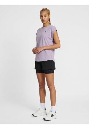 Ung kvinde med flettet hår iført en lyse lilla ærmeløs skjorte, sorte træningsshorts, hvide sokker og hvide sneakers, der står foran en ensfarvet baggrund.