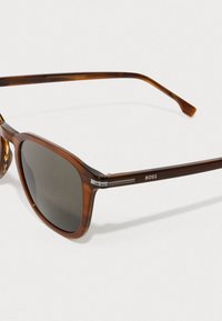 Καφέ μαρμαρωτά γυαλιά ηλίου με γκρίζους φακούς, τετράγωνο σχήμα, μεταλλικά αξεσουάρ στους κρότους και λογότυπο της μάρκας στο πλάι.