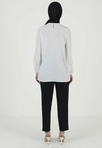 Langärmlige weiße Bluse mit einem Schlüsselloch-Rücken-Detail und elastischen Bündchen, kombiniert mit schwarzen eng anliegenden Hosen und weißen Absatzsandalen.
