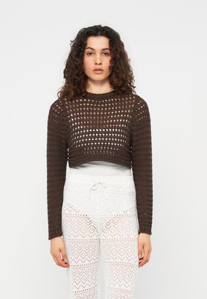 Kobieta o kręconych włosach, ubrana w ciemnobrązowy, szydełkowy, długorękawowy, krótki top na białej podkoszulce oraz białe, wzorzyste szydełkowe spodnie.