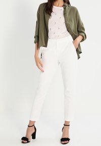 Pantaloni bianchi, camicia larga verde oliva con bottoni e una leggera maglietta sartoriale a fantasia. Sandali neri con tacco alto e dettagli con frange.