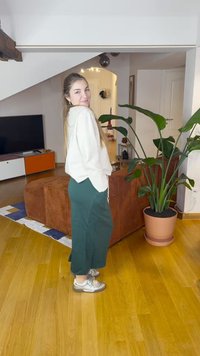 Hellfarbener, strukturierter Pullover kombiniert mit dunkelgrünen, knöchellangen Hosen. Silberne Turnschuhe und eine Topfpflanze in einem Zimmer mit Holzboden.