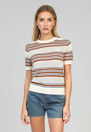 Jeune femme portant un haut tricoté à manches courtes rayé marron, bleu et blanc avec des détails œillets, assorti à un short en denim bleu.