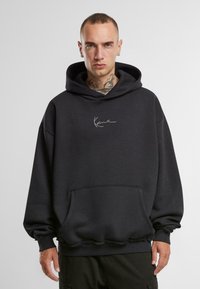 Fekete kapucnis pulóver pamutból, elöl zsebbel, bordázott mandzsettával, és fehér aláírás logóval a mellkason. Egyszerű, oversized dizájn.