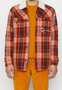 Geruite jas in rood, oranje en bruin met een witte nepbontkraag, voorzien van twee zakken en een kenmerkend detail op de borst. Draagt een oranje shirt eronder.