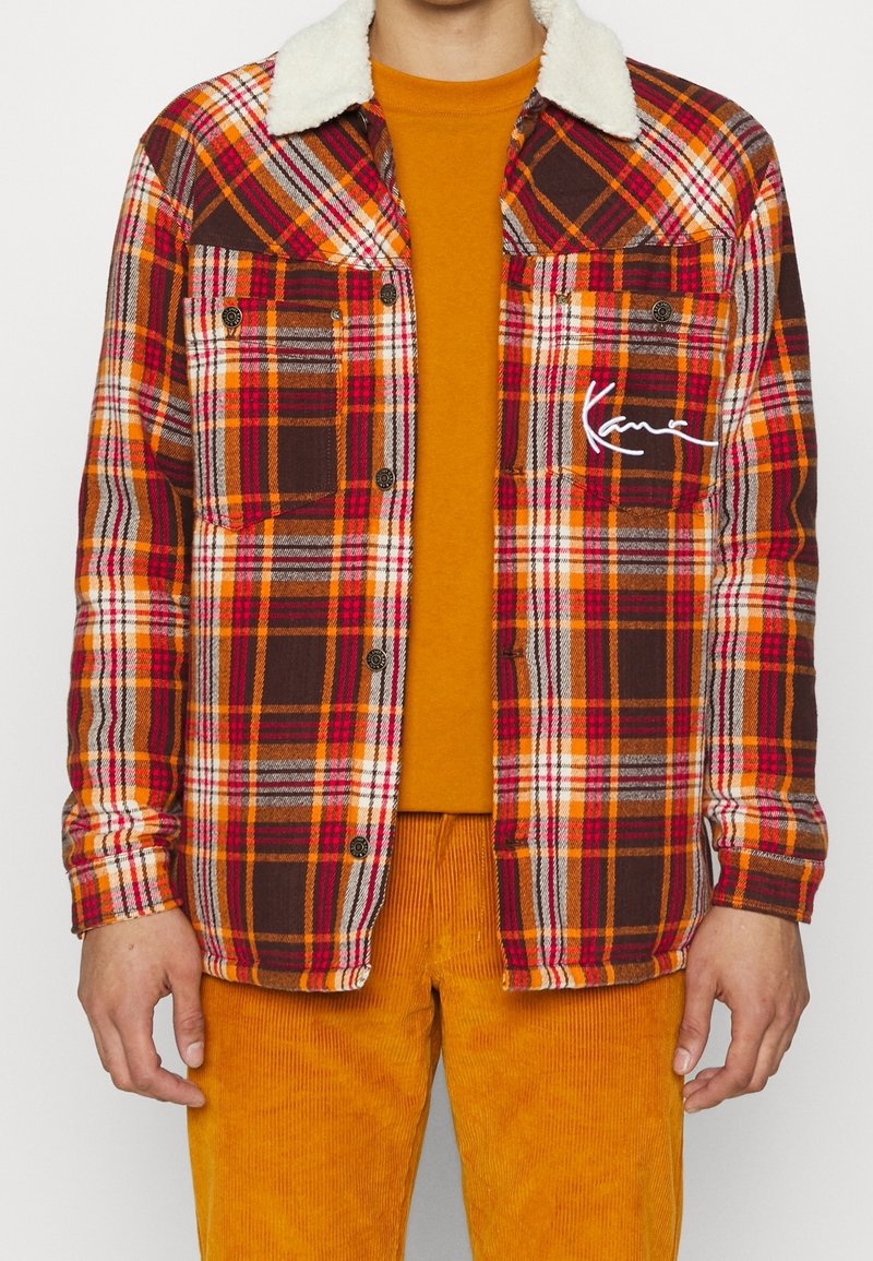 Geruite jas in rood, oranje en bruin met een witte nepbontkraag, voorzien van twee zakken en een kenmerkend detail op de borst. Draagt een oranje shirt eronder.