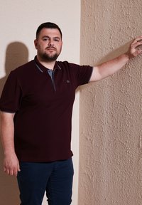 Bordový polo tričko s šedými akcenty, krátké rukávy a tříknoflíkovým zapínáním. Tmavé kalhoty. Ruce opřené o texturovanou béžovou zeď.