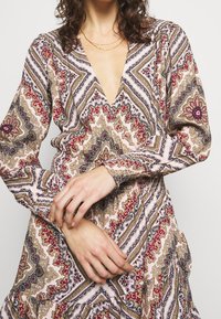El vestido multicolor cuenta con un profundo escote en V, mangas largas y un dobladillo con volantes. La tela presenta un patrón de paisley en rosa, oro y burdeos.