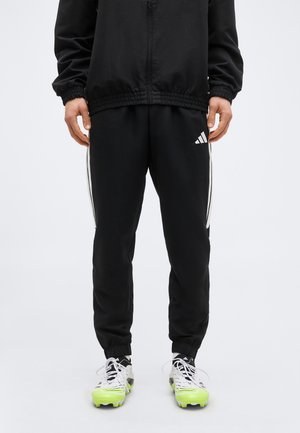 Άνδρας που φοράει μαύρο παντελόνι Adidas με λευκές ρίγες και λογότυπο, συνδυασμένο με λευκά αθλητικά παπούτσια με φωτεινές κίτρινες σόλες.