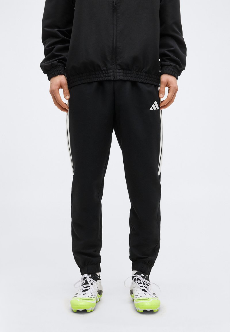 Άνδρας που φοράει μαύρο παντελόνι Adidas με λευκές ρίγες και λογότυπο, συνδυασμένο με λευκά αθλητικά παπούτσια με φωτεινές κίτρινες σόλες.