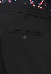 Schwarze Hosen zeichnen sich durch eine glatte Textur, eine Gesäßtasche mit einem schwarzen Knopf und einen taillierten, elastischen Bund aus.