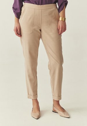 Kobieta stojąca w beżowych spodniach z mankietami, fioletowej bluzce, złotej bransoletce i beżowych balerinach na obcasie z odkrytą piętą i zakończonych spiczastym noskiem.