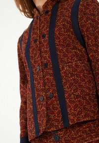 Veste tissée rouge et marine, présentant un motif texturé et de grands boutons noirs. Coupe ajustée avec des rayures contrastées sur les côtés.