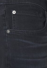 Tumma farkkujen kangas, jossa on hienovarainen tekstuuri, viiden taskun malli, metalliset napit ja kontrastinhohtoiset saumat.