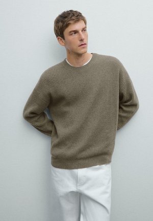 Olive green ribbed knit sweater with a relaxed fit, round neckline, and long sleeves, paired with white trousers. Soft texture and minimal design.