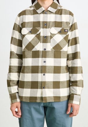 Osoba ubrana w zielono-białą, w kratę, koszulę z długim rękawem na guziki z dwoma przednimi kieszeniami z patkami oraz niebieskie spodnie, stojąca na białym tle.