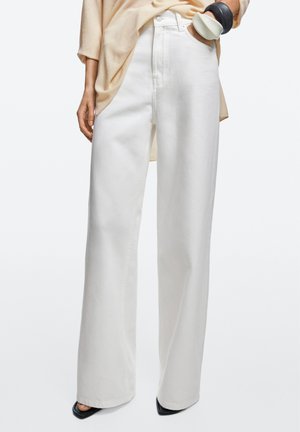 Femme portant un pantalon blanc taille haute à jambes larges, un haut beige, des chaussures noires pointues et de grands bracelets noirs et blancs au poignet.