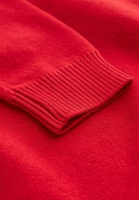 Gros plan sur la manche d'un pull rouge tricoté avec des détails de poignet côtelé sur un fond de tissu rouge assorti.