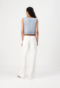Chaleco de mezclilla azul claro recortado con dobladillo sin rematar y costuras expuestas, combinado con pantalones de pierna ancha blancos de talle alto, visto desde la espalda.