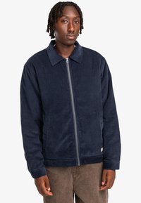 Navy corduroy zip-up jacket with a collar, side pockets, and ribbed hem. Fabric has a soft texture and a straight, relaxed fit.
