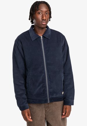 Navy corduroy zip-up jacket with a collar, side pockets, and ribbed hem. Fabric has a soft texture and a straight, relaxed fit.