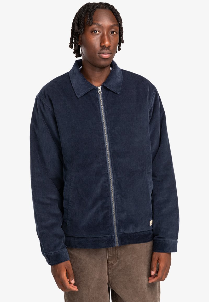 Navy corduroy zip-up jacket with a collar, side pockets, and ribbed hem. Fabric has a soft texture and a straight, relaxed fit.