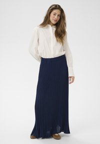 Young woman with long hair wearing a white button-up shirt, navy blue pleated maxi skirt, and brown lace-up shoes, standing against white background.