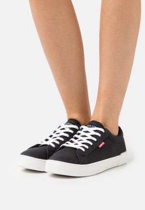 Baskets noires en toile avec des lacets blancs et des semelles en caoutchouc, arborant un logo rouge Levi's sur le côté. Design bas et look décontracté.