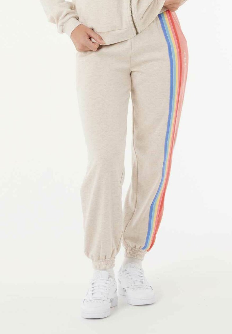 Beige sweatpants met elastische boorden, voorzien van verticale regenboogstrepen in rood, oranje, blauw en geel aan de buitenkant. Gekleed met witte sneakers.