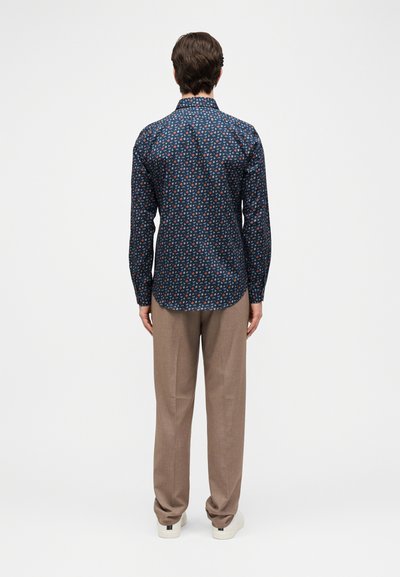 Άνδρας με την πλάτη, φοράει μπλε μακρυμάνικο πουκάμισο με σχέδια, μπεζ παντελόνι και λευκά αθλητικά παπούτσια σε λευκό φόντο.