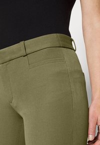 Olivgrön tight passform byxor med en slät textur, bred midjeband, bältesloopar och en enda bakficka. Närbild från höften.