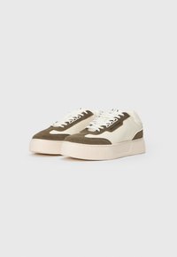 Lave sneakers med hvide såler, cremefarvede og olivengrønne stofpaneler, hvide snørebånd og et lille sort logo-mærkat på siderne.