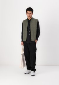 Gepolsterte olivgrüne Weste über einem schwarzen Hemd mit Knöpfen, kombiniert mit baggy schwarzen Hosen und weißen Turnschuhen, während er eine beige Tasche trägt.