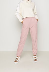 Person som bär ljusrosa joggingsbyxor med elastiska manschetter, krämfärgad kort hoodie med dragsnören och vita plattformssandaler med pärlaccen.