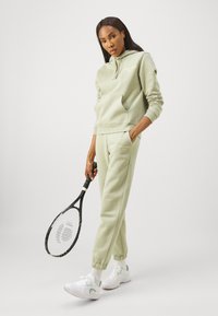 Conjunto de hoodie e calças de praia de um verde claro, com um bolso canguru, punhos canelados e logótipo bordado; combinado com ténis brancos e uma raquete de ténis.