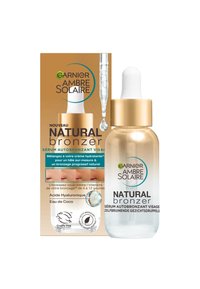 Bouteille compte-gouttes en verre transparent avec un capuchon doré, contenant un sérum clair. L'emballage présente des couleurs brunes et blanches, avec du texte et des logos bien en évidence.