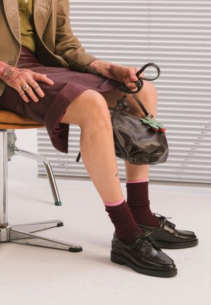 Personne assise sur une chaise de bureau tenant un sac à main en cuir noir, portant une veste marron, une jupe bordeaux, des chaussettes foncées et des chaussures noires.