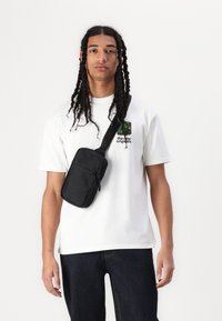 Svart crossbody-veske med en glidelåslomme foran. Har glatt tekstur og justerbar stropp, båret over en hvit skjorte og mørke jeans.
