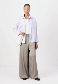 Light purple striped shirt with a patch pocket, paired with loose tan pants. Model holds a small cream bag, standing on a plain background.