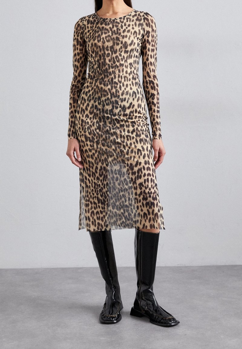 Femme portant une robe manches longues, longueur genou avec imprimé léopard, associée à des bottes noires brillantes montant jusqu'aux genoux, debout sur un sol gris.