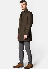 Oliwkowy wełniany płaszcz z wyciętymi klapami, dwoma kieszeniami z przodu i jednorzędowym zapięciem, zestawiony z szarymi dopasowanymi spodniami i brązowymi butami.