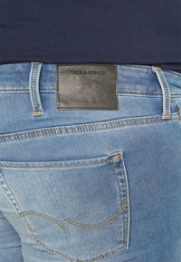 Světle modré džínové kalhoty s černou koženou etiketou. Obsahují přišité zadní kapsy s žlutými akcenty a designem obepínajícího pasu.