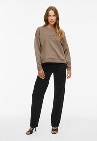 Brun sweatshirt med "TRUTH" präglad, kombinerad med svarta vida byxor och svarta klacksandaler, framför en ren vit bakgrund.